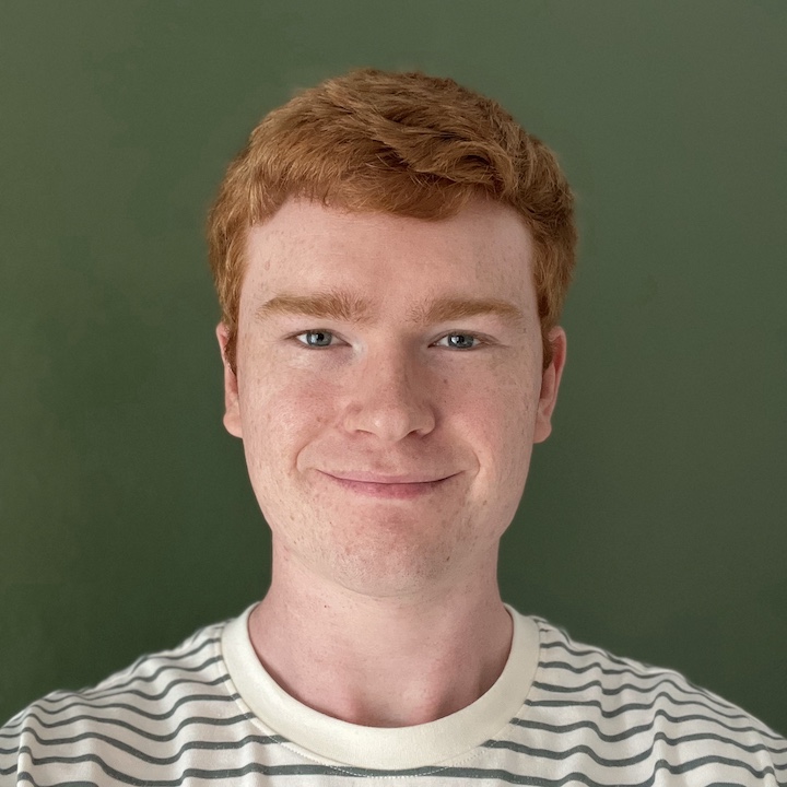 A smiling white male with short, red hair, standing in the center of the image, wearing a stripey shirt.