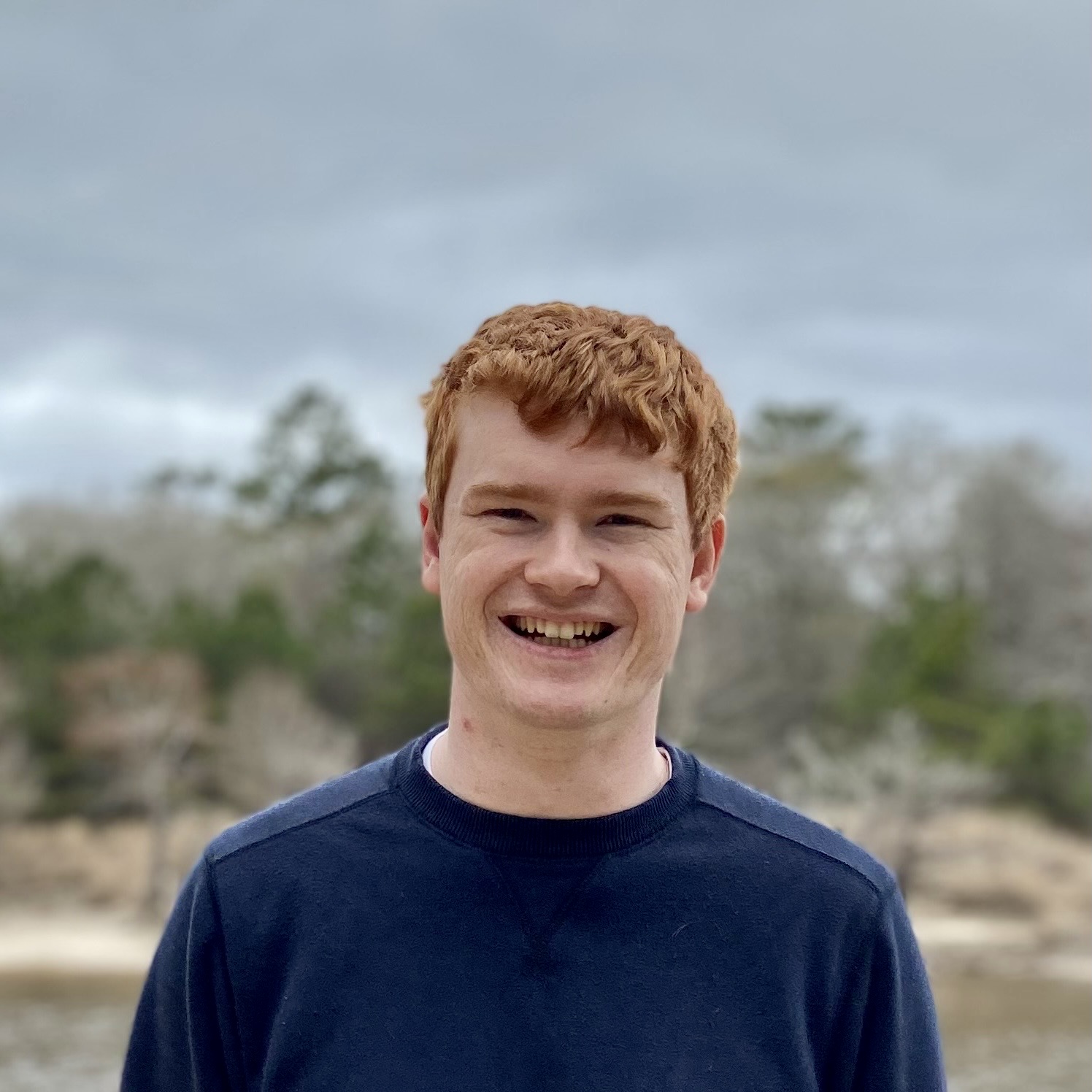 A smiling white male with short, red hair, standing in the center of the image, wearing a dark blue jumper.